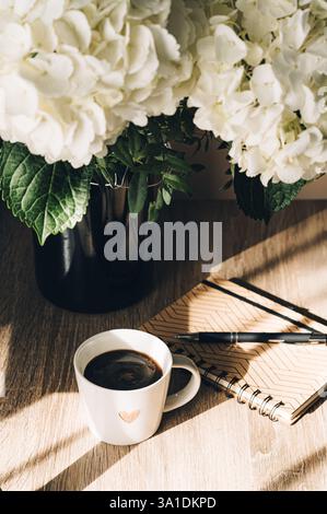 Заголовок area di lavoro con scrivania e portadocumenti in carta bianca, bouquet di fiori di ortensia blu, tazza di caffè e tastiera su sfondo bianco in legno Foto Stock