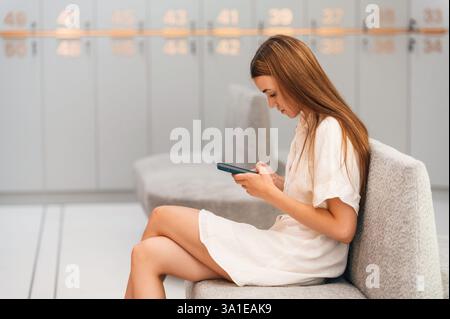 Stare seduti con lo smartphone in mano. La donna è nello spogliatoio della palestra con armadietti. Foto Stock
