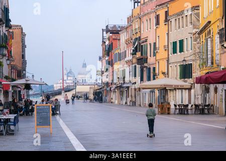 Venezia, Italia - 12 ottobre 2024: Una tranquilla mattinata su una strada veneziana sul lungomare con edifici colorati, caffetterie all'aperto e un bambino su uno scooter Foto Stock