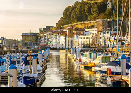 Vista panoramica del porticciolo di San Sebastián, inondata dalla luce dorata del tramonto. Le barche sono ormeggiate lungo i moli, con l'acquario sullo sfondo. Foto Stock