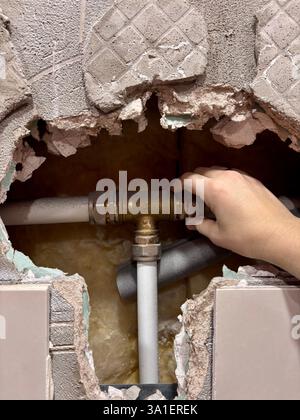 Una persona che tocca con mano un tubo in ottone all'interno di una parete danneggiata con tubazioni esposte. Le piastrelle rotte e l'intonaco sbriciolato rivelano i tubi dell'acqua e l'isolamento, indicando la riparazione domestica o la manutenzione idraulica di emergenza. La scena suggerisce problemi di perdite idriche, lavori di ristrutturazione o riparazione delle infrastrutture. Foto Stock