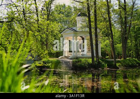 Un tranquillo parco caratterizzato da un piccolo gazebo bianco con colonne, circondato da lussureggianti alberi verdi e riflesso in un tranquillo laghetto. Foto Stock