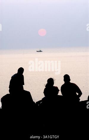 Tre turisti seduti sulle rocce e che guardano il tramonto a Harihareshwar Beach, il mare Arabico, la regione di Konkan Raigad Maharashtra Foto Stock