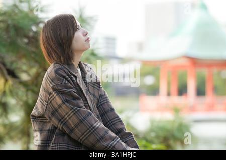 Inverno all'Ohori Park a Chuo Ward, Fukuoka City, Giappone. Una donna giapponese di 29 anni che indossa un cappotto a scacchi. Si siede su una sedia vicino allo stagno e al wat Foto Stock