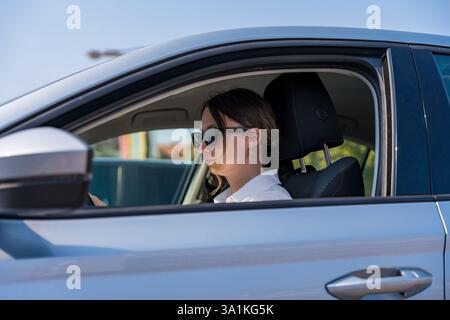 Donna caucasica che indossa occhiali da sole neri e una camicia bianca che guida un'auto d'argento in città. Foto Stock