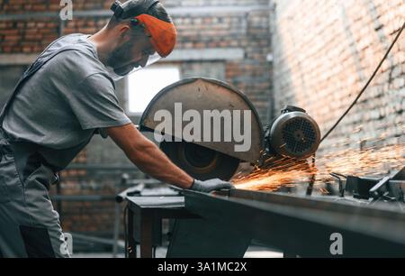 Concentrati sul lavoro, tagliando il metallo. Giovane uomo in fabbrica in uniforme grigia. Foto Stock