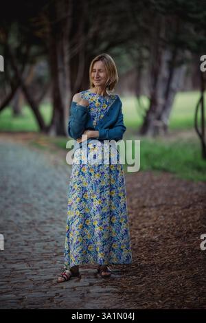 Una donna con un lungo abito estivo si erge su un sentiero di pietra in un parco durante la primavera. Foto Stock