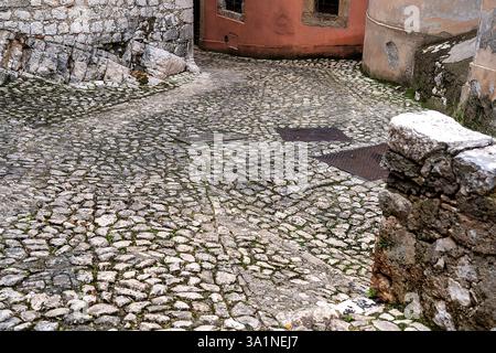 Sentiero acciottolato in discesa in un antico villaggio italiano. in fondo alla discesa un muro rosso con piccole finestre Foto Stock