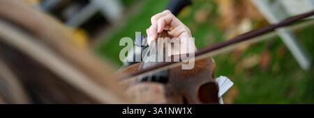 La mano di un violinista preme le corde di un violino usurato, con l'arco in movimento. Lo sfondo è sfocato e mostra un'ambientazione all'aperto con gras verde Foto Stock