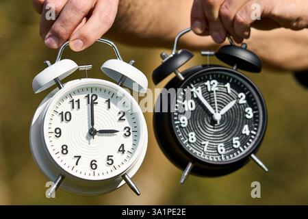 Baviera, Germania - 9 marzo 2025: Uomo con due sveglie, una che mostra l'ora legale, l'altra l'ora invernale. Tempi diversi simboleggiano il cambio di orario e il cambio di stagione *** Mann hält zwei Wecker, einer zeigt Sommerzeit, der andere Winterzeit. Unterschiedliche Uhrzeiten symbolisieren die Zeitumstellung und den Wechsel der Jahreszeiten Foto Stock