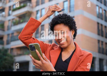 Donna sorridente che pettina i capelli e utilizza lo smartphone in uno sfondo urbano Foto Stock