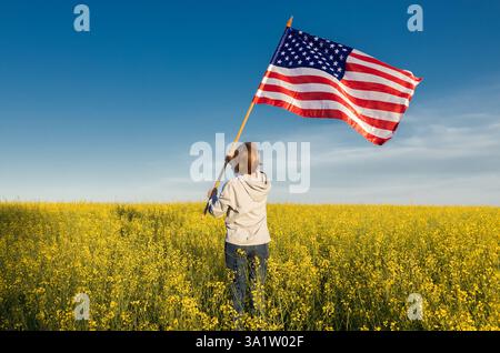 La donna sta con la schiena in un campo di colza in fiore in una giornata di sole e tiene una grande bandiera americana contro un cielo blu. Giorno di indipendenza festivo di Foto Stock