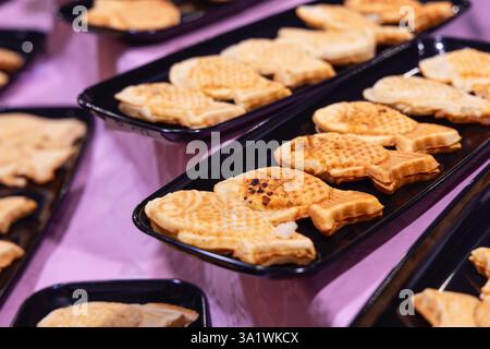 I dolci taiyaki appena sfornati a forma di pesce con una crosta croccante dorata sono disposti su vassoi neri serviti come dessert tradizionale del cibo di strada giapponese Foto Stock