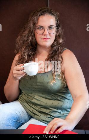Bella giovane donna con lunghi capelli ricci che tiene una tazza di caffè caldo e riposa la mano su un libro aperto, mentre si gode un momento calmo e introspettivo Foto Stock