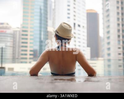 Giovane donna asiatica che si rilassa a bordo piscina all'hotel Kuala Lumpur con vista sui grattacieli circostanti, godendo del tempo libero in un vivace ambiente urbano. Foto Stock