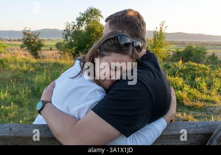 Felice e allegra coppia di mezza età seduta su una panchina in un parco che si affaccia su un campo e sulle montagne, abbracciando e sorridendo con amore. Gioia e relazione. Foto Stock
