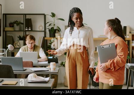 Ampia ripresa di una donna afroamericana sicura di sé che mostra la scrivania a una nuova lavoratrice in ufficio decorata con scaffali minimalisti e piante da casa Foto Stock