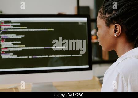 Medium close up of African American woman looking at screen with codes carefully while working in office Foto Stock