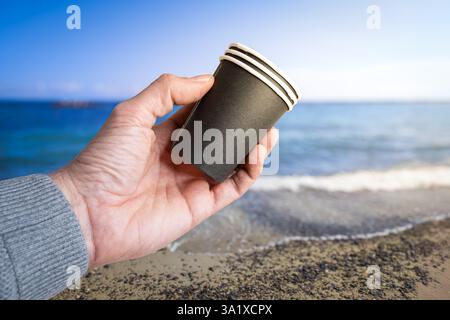 Tenere a mano tazze di plastica monouso sulla spiaggia: Concetto di inquinamento Foto Stock