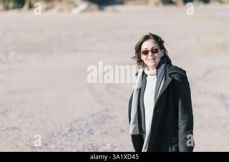 Una donna di 50 anni, che indossa un cappotto e occhiali da sole, si erge su una spiaggia illuminata dal sole, godendosi una tranquilla passeggiata invernale Foto Stock