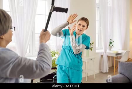 Ritratto di anziana anziana arrabbiata che colpisce la giovane infermiera con la stampella. Foto Stock