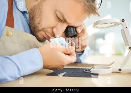 Un gioielliere mirato che esamina l'anello con lente d'ingrandimento in studio, mostrando la sua esperienza nella lavorazione artigianale. Foto Stock