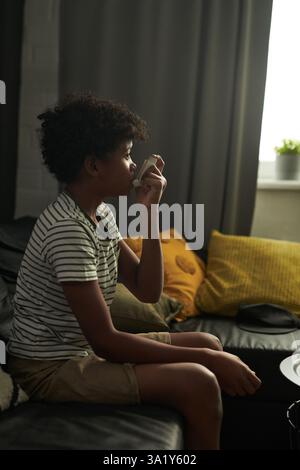 Vista laterale del giovane in un abbigliamento casual che pompa inalatore e aerosol durante l'attacco d'asma mentre è seduto sul divano Foto Stock