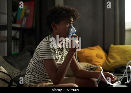 Vista laterale di un ragazzo giovane con asma che inala fumi dal nebulizzatore e guarda la tv in soggiorno mentre sei a casa Foto Stock