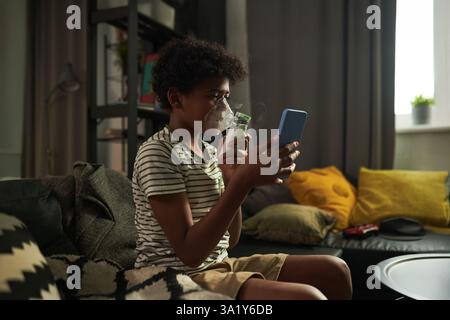 Vista laterale del ragazzo giovane seduto sul divano in soggiorno e guardare video in smartphone durante il trattamento medico di asma Foto Stock