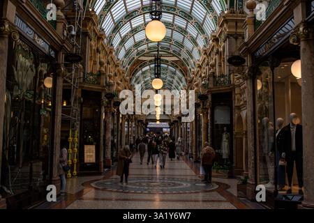Vista interna di una galleria di negozi di epoca vittoriana con persone, architettura ornata, tetto in vetro e lampade sferiche. Foto Stock