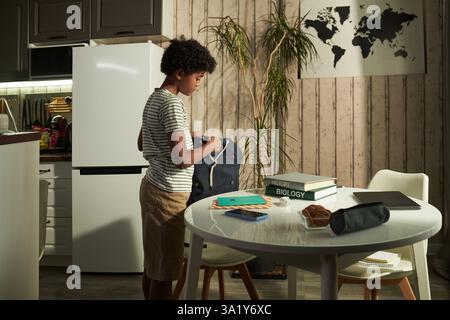 Vista laterale dello scolaro con asma che mette gli oggetti scolastici nello zaino dopo la colazione stando accanto al tavolo della cucina Foto Stock