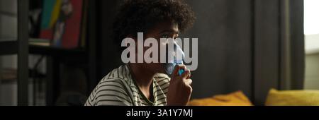 Un ragazzo giovane con asma che inala i fumi del nebulizzatore e guarda la tv in salotto mentre sei a casa Foto Stock