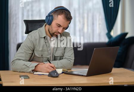 Un giovane che lavora a casa, indossa le cuffie wireless, partecipa a una videochiamata su un laptop, scrivi appunti, partecipa a un webinar di apprendimento o a un corso di formazione online. Foto Stock