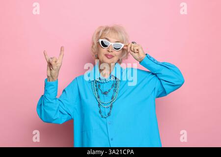 Nonna simpatica che mostra un gesto rock su sfondo rosa Foto Stock