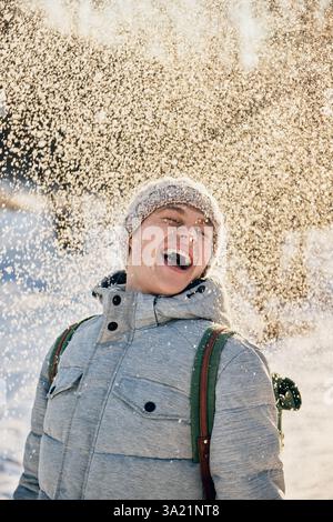 Un giovane sorridente vestito con giacca invernale grigia e cappello a maglia ama le nevicate nel soleggiato parco invernale. Foto Stock