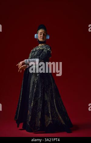 Donna anziana in abito rinascimentale in piedi con le mani in bilico, indossa cuffie futuristiche, trasuda eleganza nobile su sfondo rosso intenso. Foto Stock