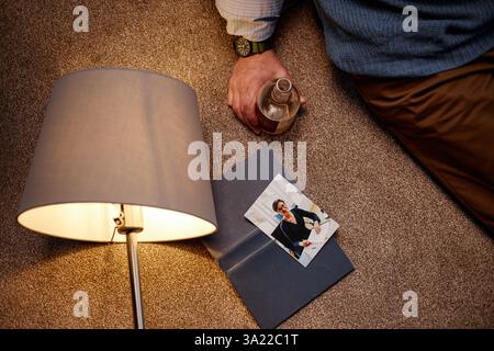 Persona seduta sul pavimento della moquette che tiene la bevanda mentre legge un documento sotto la luce calda della lampada, creando un'atmosfera tranquilla e mirata Foto Stock