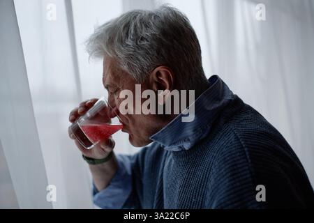 Uomo anziano con capelli grigi che beve tranquillamente bevande rosse dal vetro, in piedi accanto alla finestra in un ambiente sereno, indossa camicia blu e sembra premuroso Foto Stock