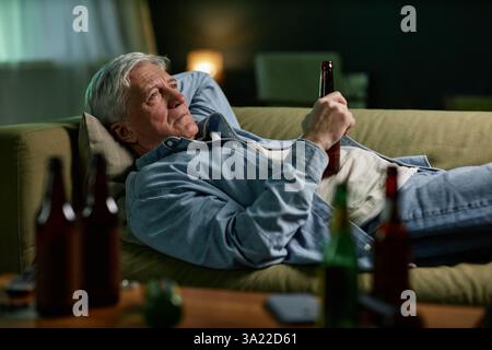 Uomo anziano che si rilassa su un divano mentre tiene un drink in una stanza poco illuminata, catturando un momento di relax e introspezione Foto Stock