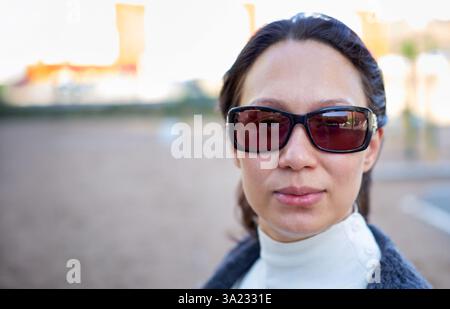 Ritratto ravvicinato di donna con eleganti occhiali da sole e abiti caldi, all'aperto con sfondo urbano sfocato. Concetto di moda e sicurezza Foto Stock