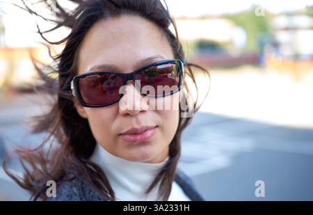 Ritratto di una donna dai capelli scuri che indossa gli occhiali da sole, i suoi capelli soffiano nel vento, in piedi all'aperto in una giornata luminosa con uno sfondo sfocato Foto Stock
