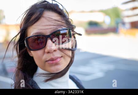 Ritratto di una donna dai capelli scuri che indossa gli occhiali da sole, i suoi capelli soffiano nel vento, in piedi all'aperto in una giornata luminosa con uno sfondo sfocato Foto Stock