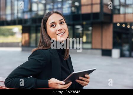Bella donna d'affari sorridente che tiene in mano un tablet digitale e guarda lontano. Giovane donna in abito formale nero seduta fuori. Foto Stock