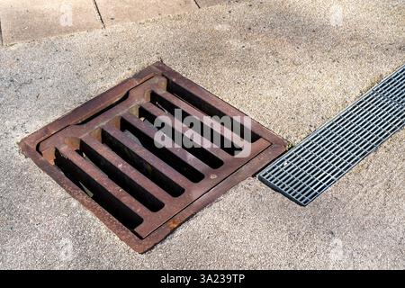 Primo piano di un vecchio coperchio di scarico in ghisa arrugginita accanto a un moderno canale di drenaggio in metallo incorporato in una superficie in cemento. Foto Stock