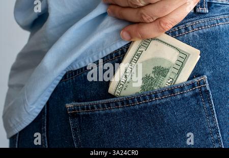 Primo piano di una persona che indossa jeans blu e camicia azzurra che mette una pila di banconote in dollari nella tasca posteriore. Concetto di risparmio, cartoline finanziarie Foto Stock