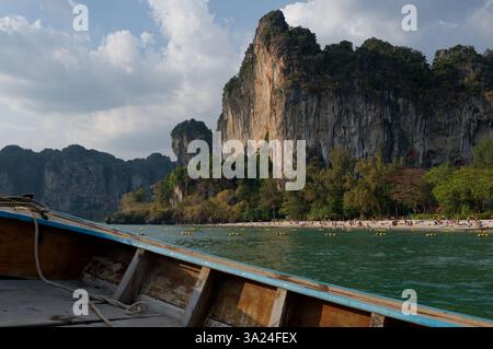 La tradizionale barca in legno si avvicina alla spiaggia di Railay, alle scogliere calcaree che sovrastano il litorale e ai visitatori Foto Stock