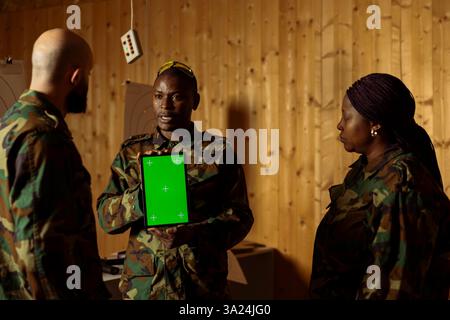 Squadra di unità militari al poligono di tiro guardando video su un tablet con schermo verde per imparare le abilità di combattimento necessarie. Soldati dell'esercito che guardano le istruzioni sul dispositivo chiave Chroma durante l'addestramento Foto Stock