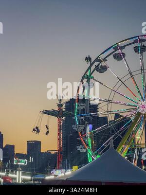 Una ruota panoramica illuminata e un giro a dondolo sullo skyline della città al crepuscolo. Il parco divertimenti presenta luci vivaci e un edificio sagomato Foto Stock