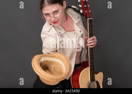 Giovane chitarrista donna con chitarra in una mano e cappello di paglia nell'altra mano allungato in avanti. Creativity Street Musician chiede donazioni per giocare Foto Stock