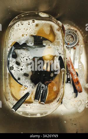 Una vista ravvicinata di un lavandino in acciaio inossidabile pieno di piatti sporchi immersi in acqua e sapone, un compito universale di lavare i piatti Foto Stock